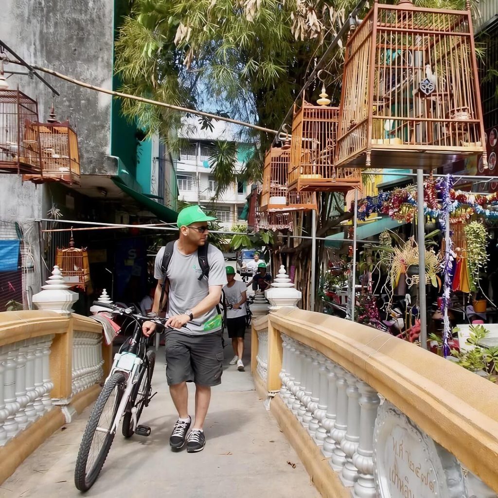 Guests pushing bicycles across a narrow footbridge with hanging bird cages in Bangkok
