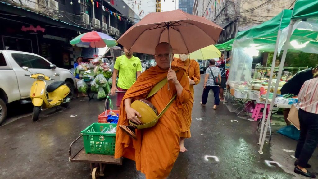 Two monks in orange robes walk through Patpong Morning Market, Bangkok. The monk in the foreground carries an alms bowl and umbrella, followed by a man with a trolley.