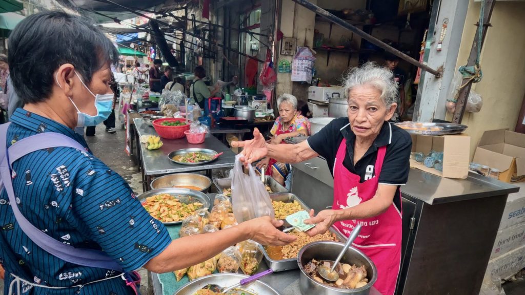 An elderly Thai woman sells traditional street food to a local customer at Trok Mor Market in Bangkok’s Old Town — a hidden gem known for its authentic, community-based culture.