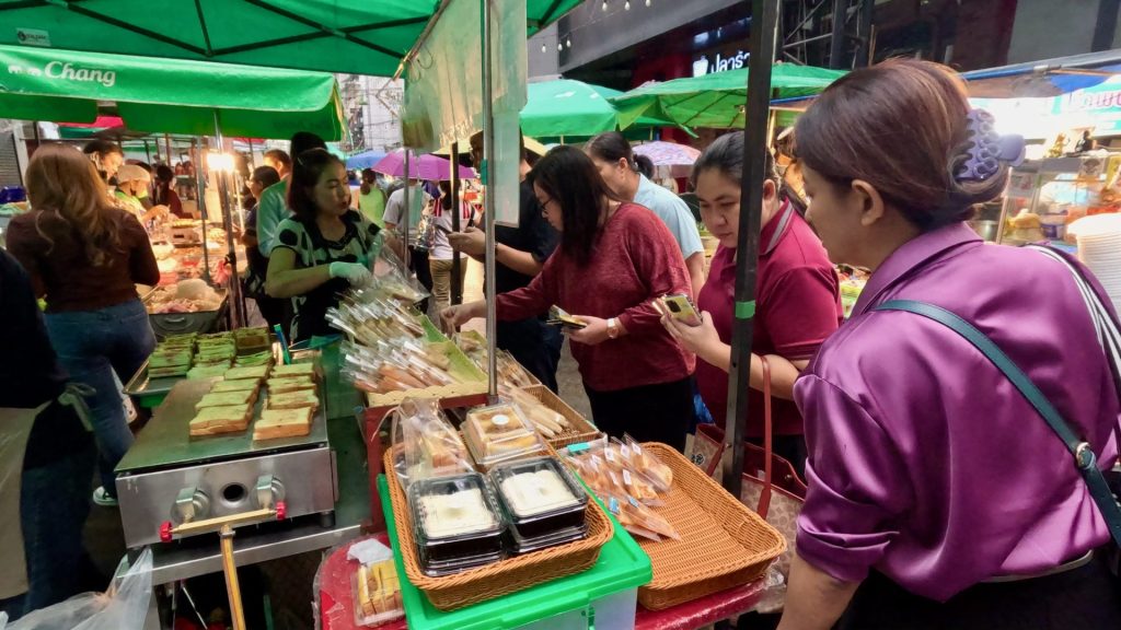 Several shoppers buying toasted sandwiches at a street food stall in Patpong Morning Market, Bangkok. Green umbrellas shade the stall, with one shopper in a silk purple top.