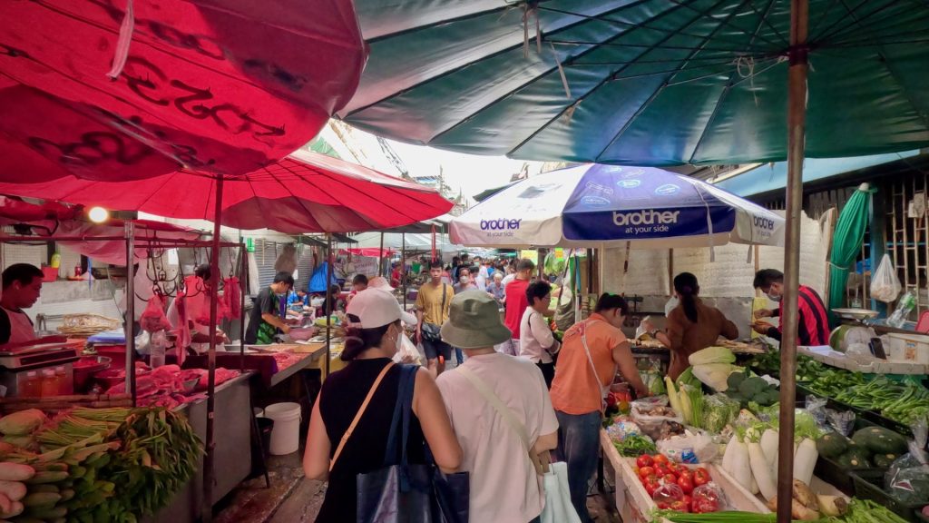 A vibrant scene at Trok Mor Market in Bangkok’s Old Town, with vendors and customers filling the narrow alleyway beneath a glowing canopy of red and green umbrellas. A slice of daily Thai life in one of the city’s most authentic local markets.