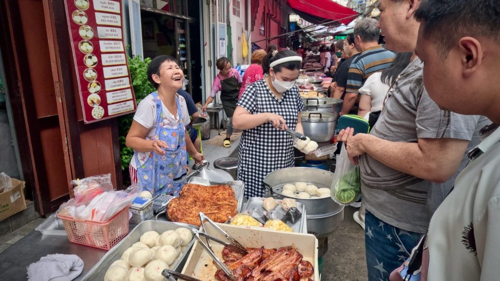 A joyful moment at Trok Mor Market in Bangkok’s Old Town, featuring an elderly female vendor laughing with a customer while serving roast pork, grilled pork skewers, and steamed dumplings at her busy street food stall.