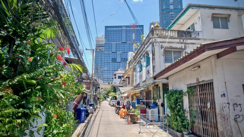 A tranquil residential alley in Bangkok with two and three-storey townhouses, clear blue sky, and the One Bangkok development in the background.