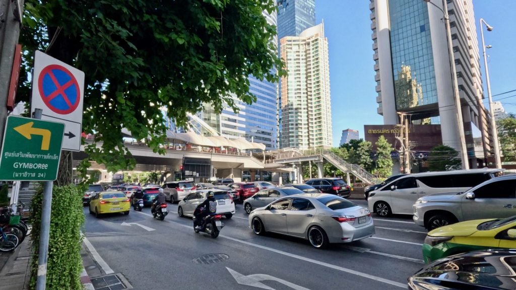Vibrant Bangkok city scene with Chong Nonsi Skywalk seen from Sathon Road, busy traffic, and modern office towers under a bright blue sky.