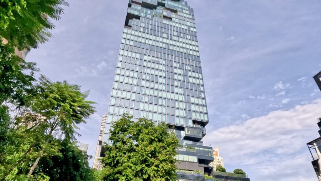 Bangkok’s Mahanakhon Tower rising above green trees, a glass-clad skyscraper set against a blue sky with fluffy white clouds, symbolising modern architecture in Thailand.