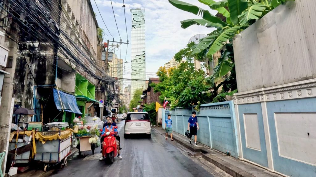 View of Bangkok’s Mahanakhon Tower from Soi Phiphat, a narrow street with pedestrians, vehicles, and a striking contrast between old townhouses and the modern skyscraper.