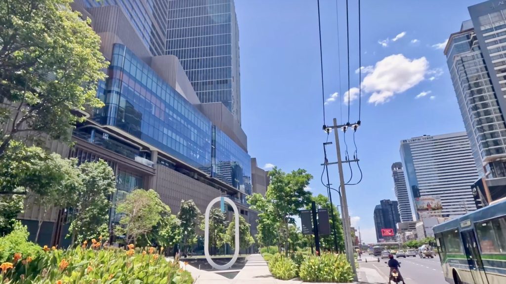 View of One Bangkok skyscrapers from Rama IV Road with landscaped gardens in the foreground and blue sky above.