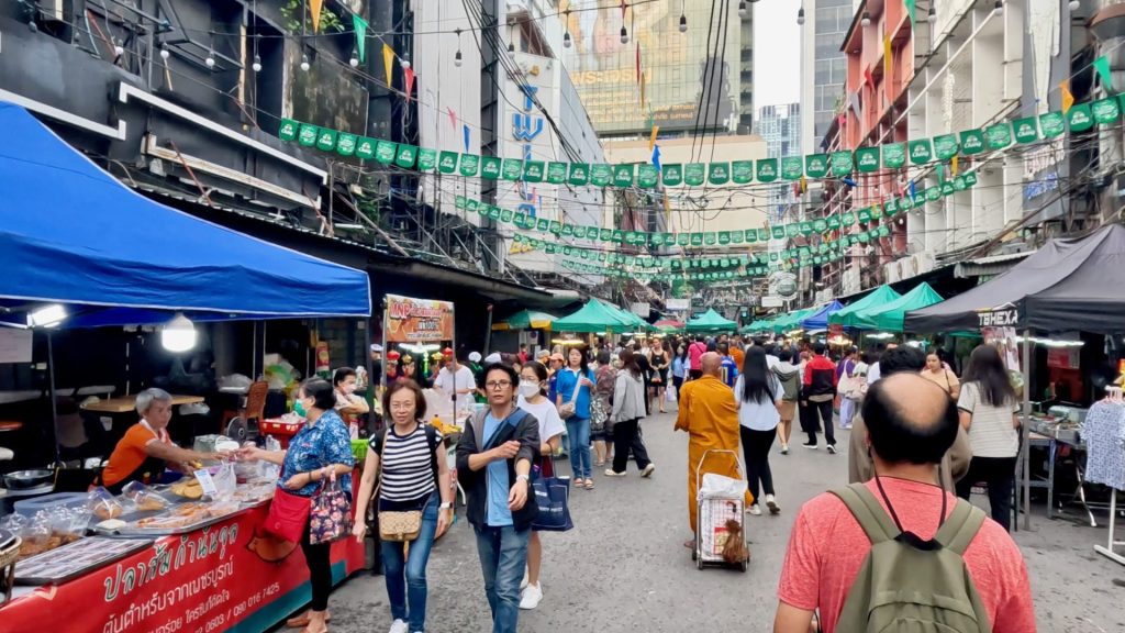 Early morning at Patpong Market in Silom, Bangkok, with monks collecting alms, green umbrellas overhead, shoppers browsing stalls, and flags strung across the busy street.