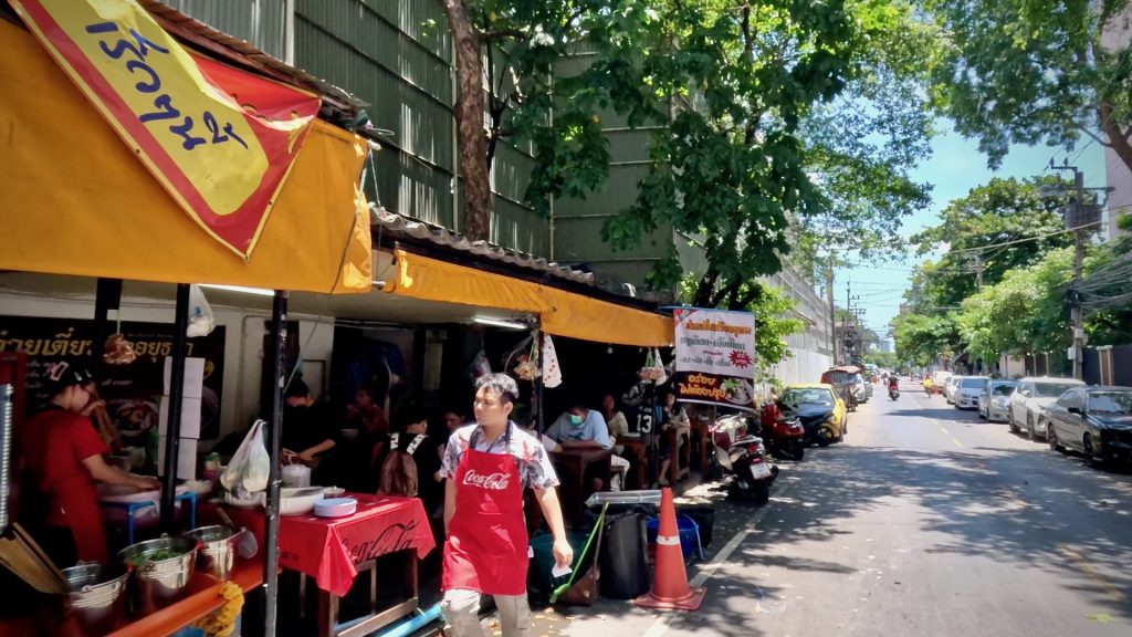 People enjoying food at a street-side restaurant on Pluk Chit Alley, a quiet street with green trees and blue sky in Bangkok.