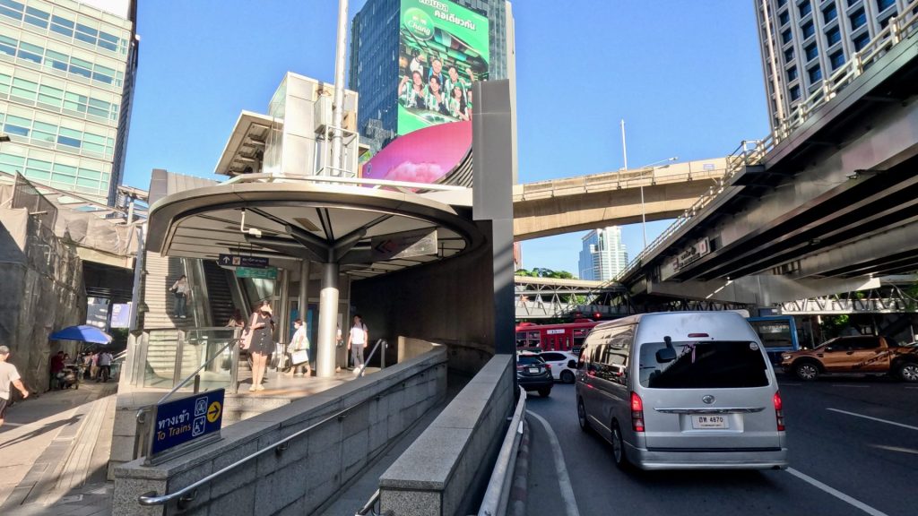 A busy morning at Silom MRT Station in Bangkok, with commuters exiting, heavy traffic on Rama IV Road, the BTS skytrain track, and tall office buildings under a clear blue sky.