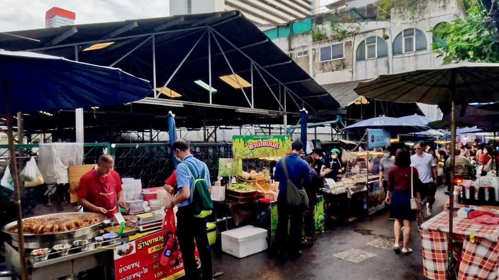 View of Silom Soi 7 morning market on a dull day in Bangkok, with vendors preparing cooked food and fresh produce, and locals shopping for daily essentials.
