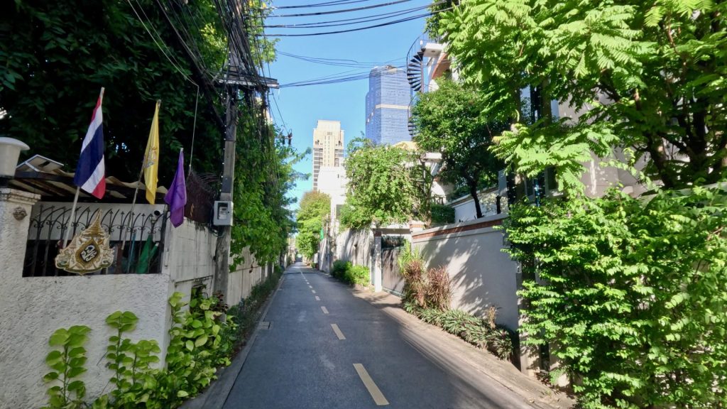 Narrow and quiet Soi Phra Phinit in Bangkok with lush greenery, shaded sidewalks, and distant office towers beneath a clear blue sky.