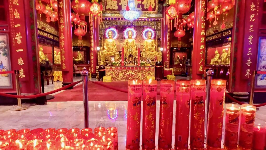 Inside Wat Mangkon Kamalawat temple in Chinatown, Bangkok, with a golden shrine, red columns, burning candles, and hanging lanterns.