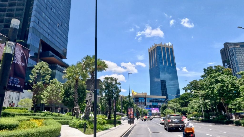 View of Witthayu Road in Bangkok with One Bangkok development on the right, a glass tower in the background, and Lumphini Park lined with green trees under a blue sky.