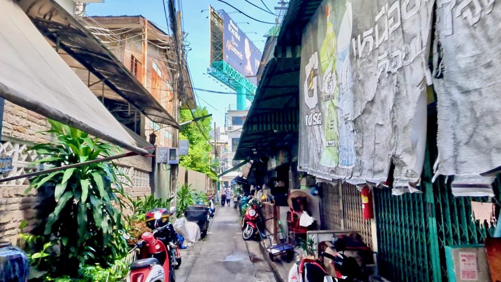 View down Charoen Rat Alley in Bangkok, a quiet alley lined with shop houses and parked motorbikes under a bright blue sky, showing local community life.