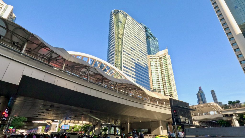 View of the Chong Nonsi Skywalk in Bangkok from Sathon Road, surrounded by glass office towers under a clear blue sky – a symbol of Bangkok’s modern cityscape.