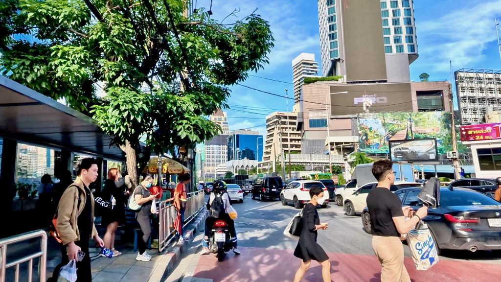 Early morning view of Sathon Road in Bangkok, showing busy commuter traffic, pedestrians crossing, and hotels and office buildings under a bright blue sky.