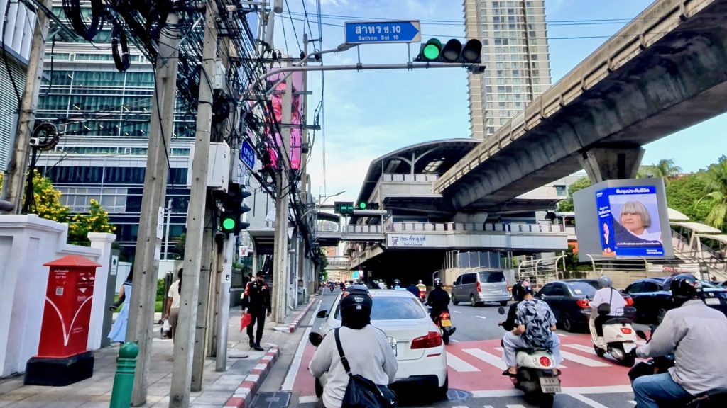 View along Sathon Road towards Saint Louis BTS Station with morning traffic, office buildings on the left, and the Skytrain track running above the road.