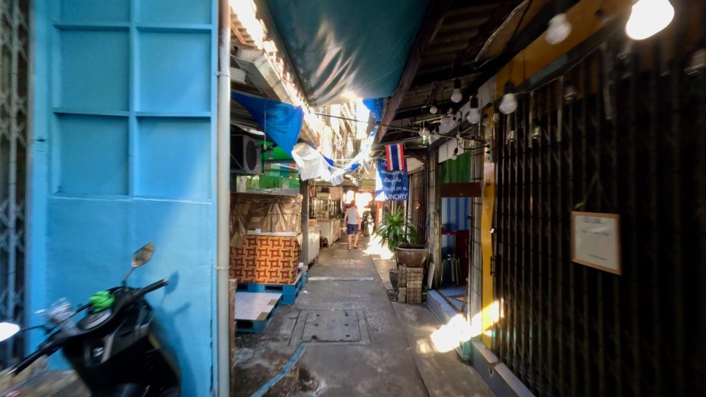 Peaceful morning view down Sathon Soi 13 in Bangkok, with sunlight filtering between awnings over small shop houses and a pedestrian walking through the narrow alley.