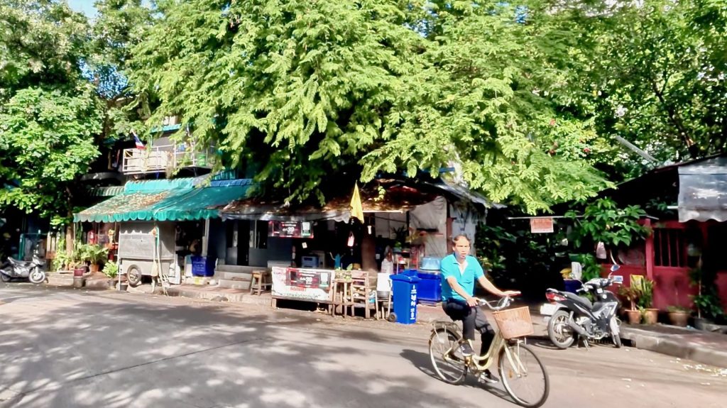 A quiet morning on Soi Ngam Duphli Alley, Sathon district, with a local man cycling past small shops shaded by green trees — showing Bangkok’s everyday neighbourhood life.