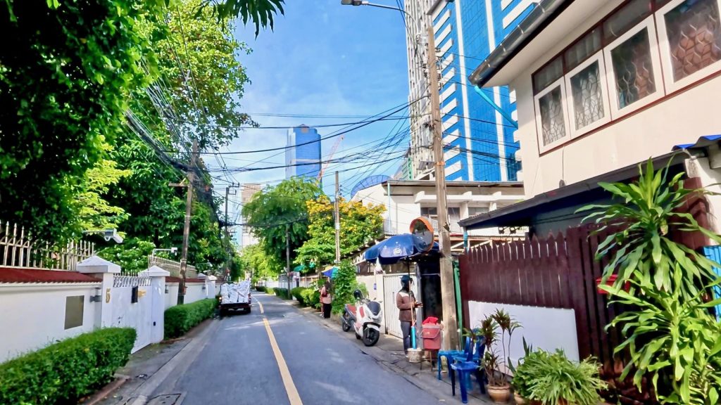 A peaceful morning on Soi Phra Phinit with greenery, local people, small homes, and walled gardens — contrasted by modern office towers in the background under a clear sky.